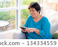 Senior asian woman smile while using on smartphone sitting on sofa in living room at home. 134334559