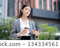 Happy young asian businesswoman standing holding coffee while using tablet outdoors city. 134334561