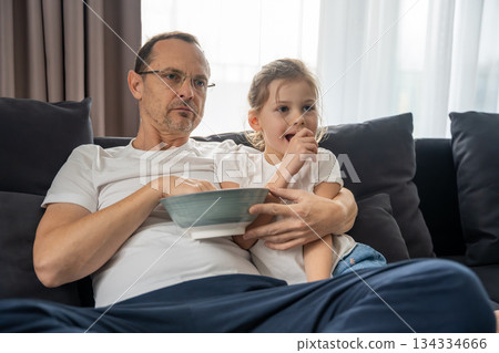Man and girl sitting on the couch eating popcorn and watching TV. Shared home activities and parent-child bonding. 134334666