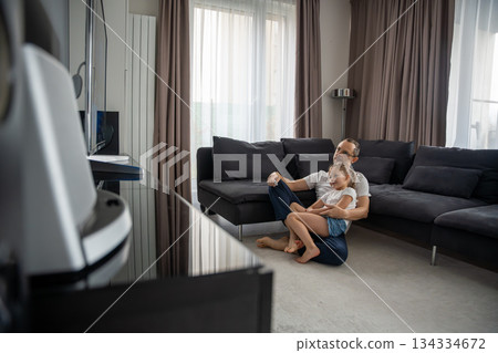 Father and daughter watching TV while sitting on the carpet in the living room. Quality family leisure time at home. 134334672
