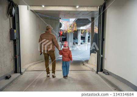 Man holding child's hand walking through garage entrance. Morning walk with child in residential area. 134334684