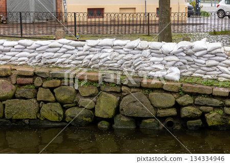 Sandbags form protective barrier riverbank reinforcing stone embankment against flood erosion. Urban waterfront shows emergency preparedness infrastructure wet surfaces reflections overcast weather Sandbags form protective barrier riverbank reinforcing stone embankment against flood erosion. Urban waterfront shows emergency preparedness infrastructure wet surfaces reflections overcast weather 134334946