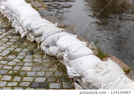 Sandbags form protective barrier riverbank reinforcing stone embankment against flood erosion. Urban waterfront shows emergency preparedness infrastructure wet surfaces reflections overcast weather 134334947
