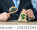 An official examines a dollar money bag with a magnifying glass. Budget assessment. Origin of capital. Financial transparency, audit procedures and investigation of funding sources. 134335244