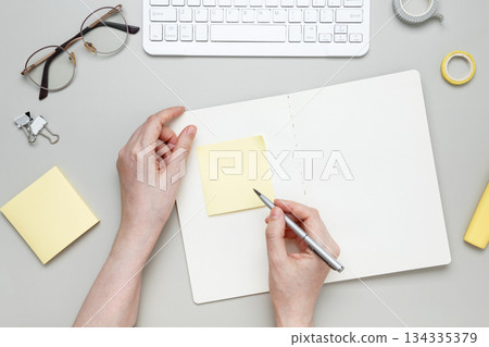 Woman writing on sticky note at organized workspace with notebook and stationery items 134335379