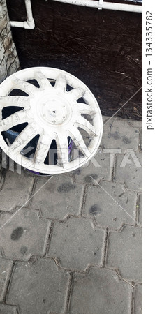 A dirty, white plastic wheel cover lies on a grey interlocking paved surface, resting against a dark wooden background and white pipes A dirty, white plastic wheel cover lies on a grey interlocking paved surface, resting against a dark wooden background and white pipes 134335782