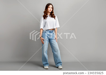 Beautiful full-length portrait of confident teenage girl with long dark hair standing in studio on gray background, looking at camera while wearing casual t-shirt and jeans 134335825