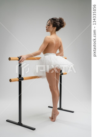 Elegant ballet dancer in nude pose with tutu by the barre in soft light 134335856