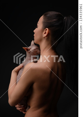 Woman embraces her hairless cat against a dark backdrop in a serene pose 134335961