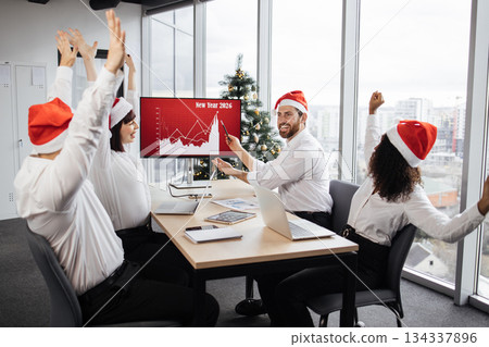 Successful team of multiethnic business people in Santa hats raising hands and screaming with happiness while working together in decorated with Christmas tree office in business center. 134337896