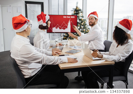 Happy team of diverse business people in Santa hats listening their middle aged male bearded chief, showing dynamics of r world market, using digital TV screen at office decorated with Christmas tree. 134337898