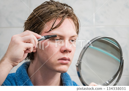 Young caucasian man using an electric trimmer to shape his eyebrows while looking into mirror in bathroom. Self-Care Routine 134338031
