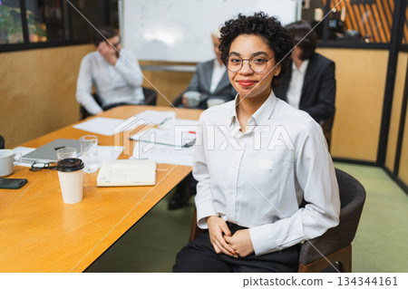 Portrait of young female team leader businesswoman in office, colleagues meeting in background. Confident African American woman business leader sitting in office. Boss leader professional worker 134344161