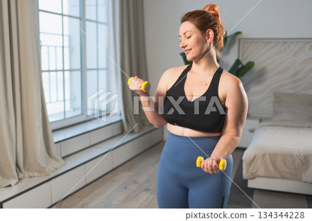 Fitness workout training. Woman picking up dumbbells. Plus size female doing sports exercise at home indoor. Girl in sportswear have workout weight training for weight loss. Sport and fitness Fitness workout training. Woman picking up dumbbells. Plus size female doing sports exercise at home indoor. Girl in sportswear have workout weight training for weight loss. Sport and fitness 134344228
