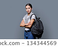 Portrait of confident female teenage student with backpack on gray background 134344569