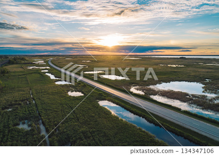 Aerial view of long rural road crossing through marshland at golden hour sunset 134347106