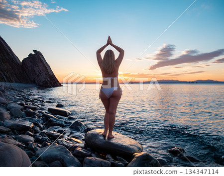 Blonde woman in a swimsuit with her back to the viewer raises her arms in a yoga pose as the sun sets behind the water. 134347114