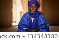 In a quiet space, a man from Mauritania sits calmly, stroking his beard as he listens closely to something. The light and shadows create a warm atmosphere around him. 134348180