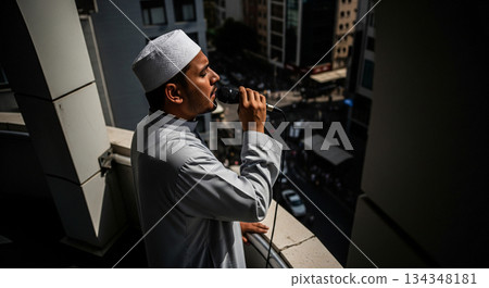 A muezzin stands at a high point, using a microphone to deliver the adhan. Below, city streets are busy as people move about, engaging in daily activities. A muezzin stands at a high point, using a microphone to deliver the adhan. Below, city streets are busy as people move about, engaging in daily activities. 134348181