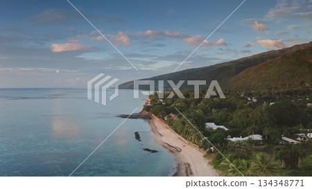 Aerial view of Tahiti island coastline featuring lush green palm trees and mountains meeting a pristine sandy beach, with calm turquoise water reflecting the sunset sky and gentle clouds. Drone flight Aerial view of Tahiti island coastline featuring lush green palm trees and mountains meeting a pristine sandy beach, with calm turquoise water reflecting the sunset sky and gentle clouds. Drone flight 134348771