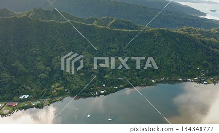 A breathtaking aerial perspective captures the vibrant green hills embracing the calm waters of a tropical bay. Sunlight reflects on the surface, highlighting the serenity of the setting. 134348772