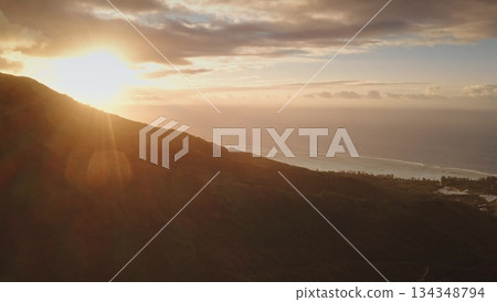 Moorea island landscape showing a vibrant sunset casting warm light over a lush green mountain slope, with the Pacific Ocean stretching towards the horizon under a soft, cloudy sky 134348794