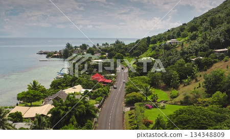 Moorea island coastal road winding through a lush tropical landscape with houses, cars, and overwater bungalows extending into the clear blue Pacific Ocean under a cloudy sky Moorea island coastal road winding through a lush tropical landscape with houses, cars, and overwater bungalows extending into the clear blue Pacific Ocean under a cloudy sky 134348809