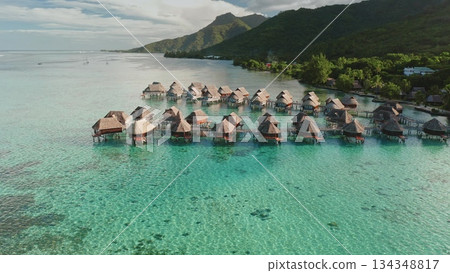 Overwater bungalows cluster across a vibrant turquoise lagoon, extending from the green tropical coastline of the French Polynesian island, capturing a luxury travel destination and tranquil scenery 134348817