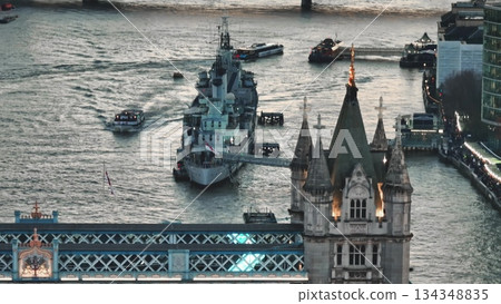England, London: HMS Belfast, Town-class light cruiser moored near Tower Bridge on River Thames, with tourist boats navigating around. Tourist attraction, famous place to visit. Aerial view 134348835