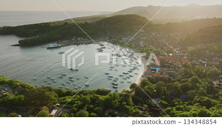 Padang Bai harbor on Bali island at sunset, fishing boats and ferries anchored in bay, coastal town nestled among lush green hills under soft light. Tropical paradise. Aerial view drone panorama 134348854