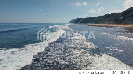 Blue ocean waves breaking on tropical sand beach with cliffs, lush green hills, people enjoying the sunny coastline of Bali island, perfect for travel and holiday concept. Aerial view drone flight Blue ocean waves breaking on tropical sand beach with cliffs, lush green hills, people enjoying the sunny coastline of Bali island, perfect for travel and holiday concept. Aerial view drone flight 134348868