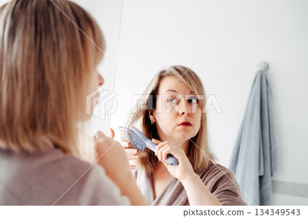 Woman looks in bathroom mirror and adjusts her wet hair with her hand. Authentic beauty, realistic portrayal, unposed moments, everyday life. 134349543