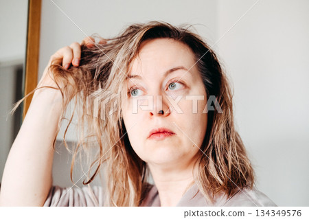 An adult woman touches her damp, wavy hair after washing it, looking up and away with a pensive expression. Scalp health, personalized haircare, wet hair routine. An adult woman touches her damp, wavy hair after washing it, looking up and away with a pensive expression. Scalp health, personalized haircare, wet hair routine. 134349576