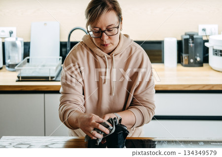 A woman wearing glasses and a hoodie wipes a black kitchen appliance on the counter with a dark reusable cloth. Eco friendly kitchen, reusable cloths, sustainable cleaning products, domestic hygiene. A woman wearing glasses and a hoodie wipes a black kitchen appliance on the counter with a dark reusable cloth. Eco friendly kitchen, reusable cloths, sustainable cleaning products, domestic hygiene. 134349579