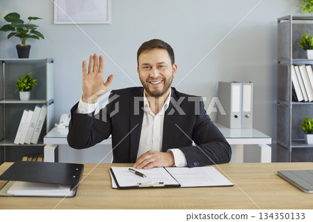 Smiling businessman at office desk waving hand, greeting employees at online business conference 134350133