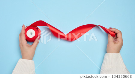 A woman's hands, wearing a white knitted sweater, hold a skein of red satin ribbon and the unfolded edge of the ribbon. A woman's hands, wearing a white knitted sweater, hold a skein of red satin ribbon and the unfolded edge of the ribbon. 134350668