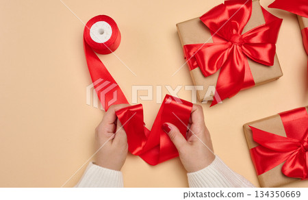 A woman's hands in a white sweater hold a gift box wrapped in craft paper with a red satin bow and a roll of ribbon. Beige background. 134350669