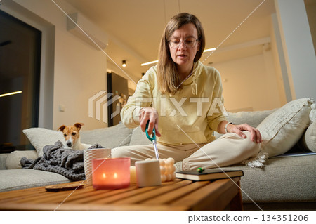 Woman sitting on sofa cutting candle wick on coffee table while small dog rests nearby 134351206