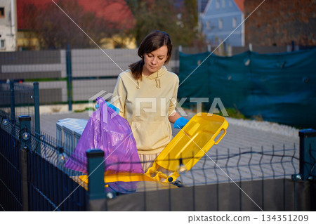 Woman wearing gloves placing garbage bag into outdoor recycling container 134351209
