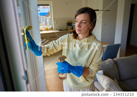 Woman wearing gloves cleaning glass window with cloth and spray bottle Woman wearing gloves cleaning glass window with cloth and spray bottle 134351210