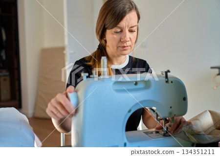 Woman sitting indoors operating sewing machine and guiding fabric 134351211