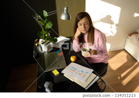 Woman sitting at home desk holding mug next to laptop, documents and office supplies 134351213