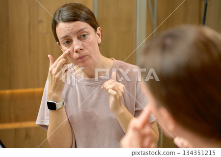 Woman standing in bathroom applying cream under eye Woman standing in bathroom applying cream under eye 134351215