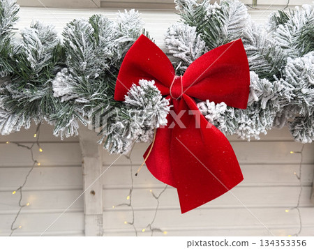Red velvet bow on snowy evergreen garland outdoors. Holiday decor detail, winter atmosphere, and classic Christmas styling 134353356