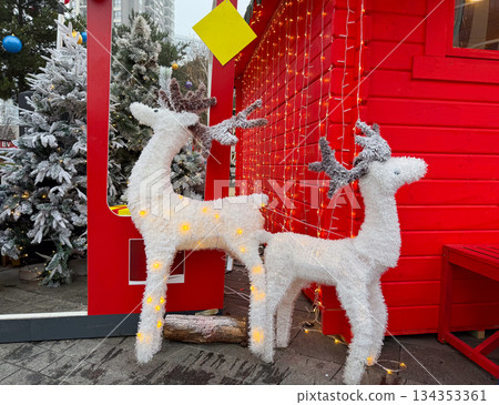 Illuminated Christmas reindeer figures near red wooden holiday house. Festive outdoor decoration, winter celebration, seasonal city market atmosphere 134353361