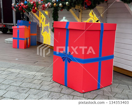 Large red gift box at christmas market stall. Festive installation, winter holiday decor, and seasonal street atmosphere. 134353403