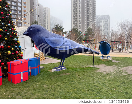 Large blue bird sculpture standing near gift boxes in winter park. Festive urban decor, public art, and holiday atmosphere 134353416