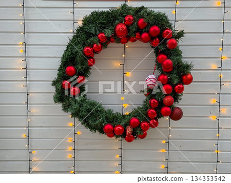 Christmas wreath with red ornaments hanging on white wall with lights. Festive holiday decor, winter atmosphere, and seasonal celebration Christmas wreath with red ornaments hanging on white wall with lights. Festive holiday decor, winter atmosphere, and seasonal celebration 134353542
