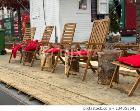 Wooden lounge chairs with red blankets near an outdoor winter ice rink. Cozy rest area, seasonal comfort, and festive urban leisure Wooden lounge chairs with red blankets near an outdoor winter ice rink. Cozy rest area, seasonal comfort, and festive urban leisure 134353543