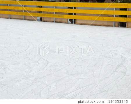 Empty outdoor ice rink with skate marks and yellow wooden barrier. Winter sports background, cold season atmosphere, and urban leisure space Empty outdoor ice rink with skate marks and yellow wooden barrier. Winter sports background, cold season atmosphere, and urban leisure space 134353545